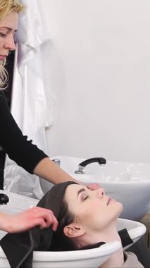 Modern bir güzellik salonunda, yetenekli bir güzellik uzmanı mutlu genç bir kadının saçlarını yumuşacık bir havluyla nazikçe yıkar. Bu sakin sahne rahatlama ve hoşgörü yayar, rahatlatıcı su akışını ve güzel kokulu şampuanı vurgular,