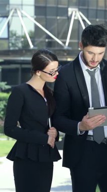 Üç dinamik iş ortağı, şık takım elbiseli kendine güvenen bir adam ve iki şık kadın, açık hava tabletindeki heyecan verici haberlere sevinçle tepki veriyorlar. Işıltılı gülümsemeleri ve hareketleriyle, çerçevelenmiş canlı bir başarı ve işbirliği atmosferini yansıtıyorlar.