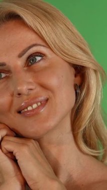 Işıl ışıl sarı saçları ve ışıl ışıl mavi gözleri olan ışıl parlayan olgun bir kadın kameraya neşeyle bakar, elleri nazikçe yüzünü canlı yeşil renkli bir arka plana yaslar. Bu büyüleyici görüntü, kadınlığın ve benliğin özünü yakalar.