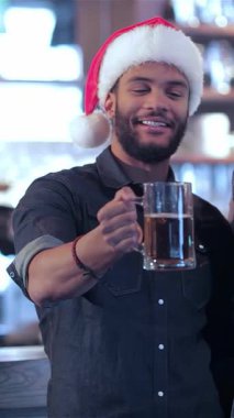 Canlı bir bar sahnesinde, iki arkadaş Noel Baba şapkalarını coşkuyla tokuştururken, bir barmen bayram neşesini paylaşıyor. Hava kahkaha ve heyecanla dolu. Sıcak bir atmosfer yaratıyor.