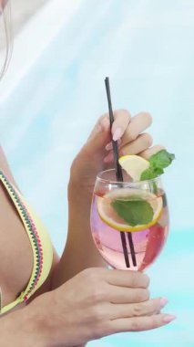 Bu canlı kırpılmış çekim renkli bikinili muhteşem bir kadını canlandırıyor, narenciye ve nane ile süslenmiş ferahlatıcı bir kokteylle zevkini çıkarıyor. Köpüklü havuz suyu güneş ışığında parlar ve yaz için mükemmel bir atmosfer yaratır.