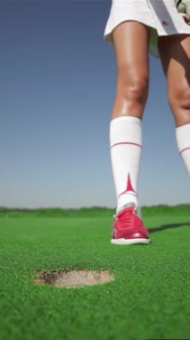 Kırmızı golf ayakkabılarına odaklanmış genç bir kadın topu fincana doğru atarken kararlılığını belli bir vuruşla ustalıkla ortaya koyuyor. Bereketli yeşil golf sahası, parlak güneş ışığı altında parıldıyor.