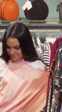 İki modaya uygun genç kadın, biri göz kamaştırıcı bir şeftali elbiseye hayran kalırken, diğeri ise yakınlarda hareketli bir tarzda geziniyor. Butikler şık dekorlar ve renkli elbiseler davetkar bir atmosfer yaratıyor.