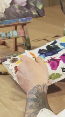 Bu samimi yakın planda, dövmeli bir erkek ressam paletine canlı yağlı boya sıkar ve yaratıcılığını rahat bir sanat stüdyosunda sergilemeye hazırdır. Bir dizi fırça ve boş bir tuval ile çevrili sahne,