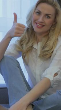 Dalgalı sarı saçlı neşeli genç bir kadın, şık, gri bir kanepede rahatça otururken parlak bir gülümseme saçar. Samimi oturma odası, doğal ışıkla yıkanmış, sıcaklığı ve pozitifliği yayıyor, izleyicileri neşeye davet ediyor.