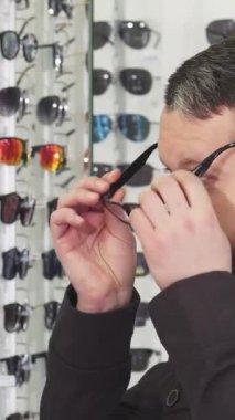 Sakallı, olgun bir adam şık bir gözlükçüdeki yansımasına bakarken moda gözlüklerini incelikle ayarlıyor. Moda çerçevelerinin göz kamaştırıcı bir dizisi ile çevrelenmiş olan yüzü heyecanla parıldıyor.