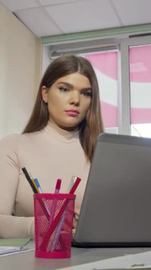 Uzun kahverengi saçlı ve sarı tırnaklı yorgun bir kız öğrenci bilgisayarına yoğun bir şekilde odaklanırken şakaklarını gıcık ediyor. Parlak, modern bir çalışma alanındaki canlı kırtasiyelerle çevrelenmiş bu resim, güçlü bir şekilde duygusal bir şekilde aktarım yapıyor.