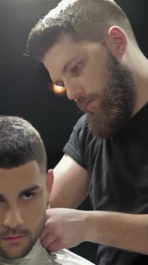 Şık bir berber dükkanında, şık sakallı uzman bir berber genç bir erkek müşteri için mükemmel saç kesimi ve sakal kesimi üzerine odaklanır. Enerjik atmosfer enerji ile uğulduyor, modern tımarcılığın ustalığını ve hassasiyetini vurguluyor.