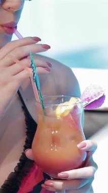 Uzun, dalgalı kahverengi saçlı büyüleyici bir beyaz kadın, ışıl ışıl havuz alanına göz gezdirerek canlı bir kokteylin tadını çıkarıyor. Şık güneş gözlükleri ve ışıl ışıl gülüşü yazın özünü temsil ediyor.