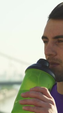 Yakışıklı bir delikanlının, yoğun bir idmandan sonra canlı yeşil bir su şişesiyle neşeyle su içtiği yakın plan bir fotoğraf. Işıl ışıl gülüşü ve odaklanmış ifadesi spor ve refahı yansıtıyor, etkin bir ruhun özünü yakalıyor.