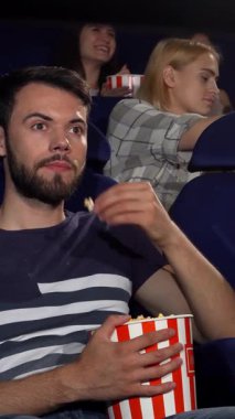 Neşeli gülümseyen genç bir adam, sinemadaki heyecan verici deneyime tamamen dalmış bir şekilde başparmağını kaldırır. Etrafı arkadaşlarıyla çevrili, klasik çizgili patlamış mısır kovasını kavrıyor, neşe ve arkadaşlığın özünü yakalıyor.