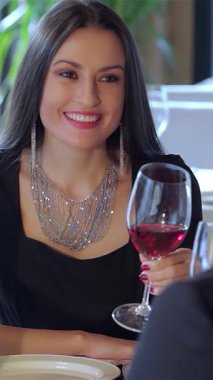 Parlak genç bir kadın, şık bir restoranda partneriyle romantik bir akşam yemeği yerken kırmızı şarap kadehini kaldırırken neşeyle ışıldıyor. Yumuşak ışıklandırma ve yemyeşil ile süslenmiş samimi ortam, onların sihirli bir zemin oluşturur.