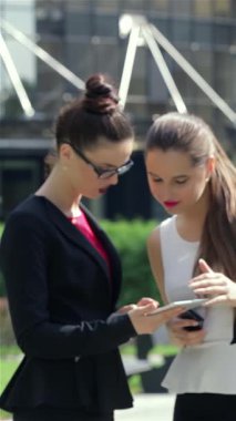 Hareketli bir şehir manzarasında, şık siyah ceketli ve şık kırmızı bluzlu kendine güvenen genç bir iş kadını tabletiyle meşgul olurken, bayan meslektaşları da yakınlarda iş birliği yapıyor. Bu canlı sahne profesyonelliği ve hırsı ön plana çıkarıyor.