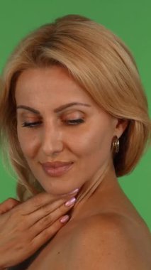 Çekici, olgun, dalgalı sarı saçlı bir kadın zarif bir şekilde kameraya doğru döner, eli parlak tenini canlı yeşil bir krom arka planda nazikçe okşar. Bu büyüleyici stüdyo portresi özgüven ve şehvet yayar.