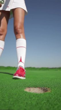 Yakından çekilen bir fotoğraf, şık bir beyaz etek ve tişört giymiş, golf topuna canlı yeşil bir sahada vurmaya hazır, kararlı, beyaz bir kadın yakalıyor. Parlak bir salıncağa hazırlanırken atletizmi parlıyor