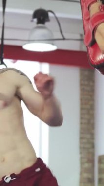 Güçlü, üstsüz bir erkek kickboksör antrenörünün tuttuğu pedlere tekme atarken atletizmini gösteriyor. Enerjik spor salonu atmosferi enerji ve kararlılıkla uğulduyor.
