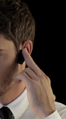 Gıcır gıcır beyaz gömlekli ve parlak siyah kravatlı ciddi bir beyaz adam kulaklığından dikkatlice dinliyor, eli kulağına yakın duruyor, nişanını vurguluyor. Dramatik siyah arkaplan, onun odaklanmış ifadesini güçlendirir.