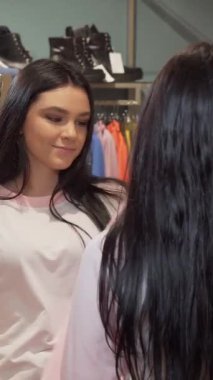 Kendine güven ve heyecanı somutlaştıran, aynanın önündeki şık pembe kıyafete hayran olan, uzun siyah saçlı şık bir genç kadın. Moda ürünleri ve hafif ışıklandırma ile süslenmiş canlı giyim mağazası, davetkar bir davet yaratıyor.