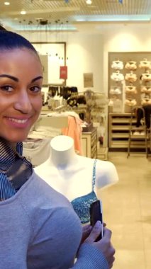 Zarif bir Afrikalı Amerikalı kadın modern bir butikte şık bir iç çamaşırı sergisinin fotoğrafını çekerken kendine güvenle ışıldıyor. Akıllı telefonuyla, bir mankenin yanında duruyor, bu canlı perakendecilikte moda tutkusu ve neşe saçıyor.