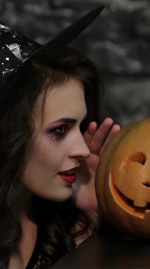 Büyüleyici bir kadın büyücü, sırıtan bir balkabağına doğru eğiliyor, parmağı kıvrımlı, siyah tuğla bir duvara karşı şakacı bir şşş hareketiyle kalkıyor. Bu Cadılar Bayramı 'ndan esinlenilmiş sahne bir gizem ve sihir havası yayıyor.