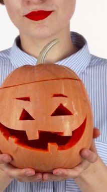 Kalın kırmızı dudaklı ışıl ışıl bir kadın neşeli bir şekilde Cadılar Bayramı 'nda oyulmuş bir balkabağı sunar, neşeli gülüşü şenlikli bir ruhla parlar. Bu canlı sahne, sonbahar kutlamalarının sihrini özetliyor. Bulaşıcı mutluluğunu vurguluyor.