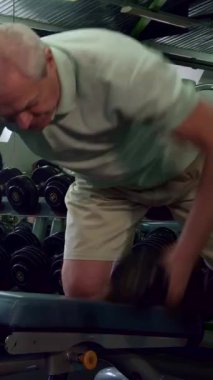 Kendini adamış genç bir Kafkasyalı antrenör, üst düzey bir erkek müşterinin spor salonunda güç egzersizleri yapmasını dikkatle izliyor ve destekleyici ve motive edici bir atmosfer yaratıyor. Sakallı fitness uzmanı yaşlı adamın yanında diz çöker.