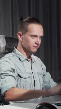 Kendini işine adamış, gündelik açık mavi gömlekli genç bir adam, modern bir masada oturuyor, gece geç saatlerde bilgisayar işleriyle meşgul, sakin bir ambiyansla çevrili. Çevresindeki ışığın yumuşak parıltısı, onun konsantre yüz ifadesini vurguluyor.