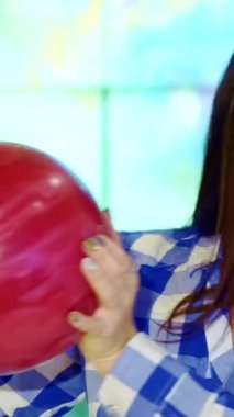 Neşeli esmer bir kız yüzüne çarpan pembe bir bovling topunu neşe ve coşku saçarak şakacı bir şekilde beşiğinde tutuyor. Bu yakın plan çekimde, şık mavi ve beyaz kareli gömlekli neşeli beyaz bir kadın, eğlence dolu bir gömlek için mükemmel hazırlanmış,