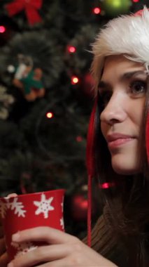 Noel Baba şapkası takmış neşeli, genç, beyaz bir kadın sıcak kakaonun, sıcaklığın ve tatil ruhunun tadını çıkarıyor. Yanıp sönen ışıklar ve Noel ağacındaki canlı süsler arasında, düşünceli bir şekilde bakıyor,