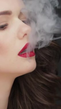 Bu büyüleyici ağır çekimde, dalgalı esmer saçlı çarpıcı beyaz bir kadın izleyiciyi cezbediyor, parlak cildi koyu siyah bir arka planda parlıyor. Girdap gibi dönen dumanın rahatsız edici varlığı onun güzelliğine tamamen zıt.