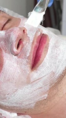 Rahat ve mutlu yakışıklı bir adam gözleri kapalı rahatça yatar. Yetenekli bir kozmetolog nazikçe rahatlatıcı bir yüz maskesi takar. Huzurlu spa ortamı deneyimi güçlendirir, cildine sağlık ve gençleşme sağlar.