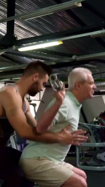 Dinamik bir spor salonu ortamında, odaklanmış genç bir erkek antrenör çömelme ve güvenliği sağlamak için kıdemli bir müşteriye destek olur. İyi bakımlı sakallı kendini işine adamış antrenör yaşlı adamın kollarını tutuyor ve güven duygusu besliyor.