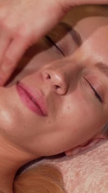 Işıl ışıl genç bir kadın gözleri kapalı rahatlar, yetenekli bir masöz huzurlu bir güzellik salonunda rahatlatıcı bir yüz masajı yaparken mutlulukla gülümser. Yumuşak ışıklandırma ve nazik dokunuş cildini geliştirmek için sakin bir atmosfer yaratıyor.