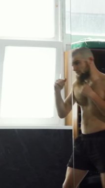 Dinamik bir çekim, iyi aydınlatılmış bir spor salonunda güçlü yumruklar atan üstsüz bir erkek MMA dövüşçüsünü yakalar ve aynadaki kararlılığını yansıtır. Bu sahne gücün, çevikliğin ve acımasız motivasyonun özünü temsil ediyor.