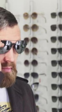 Sakallı, olgun bir adamın canlı bir gözlük mağazasında şık güneş gözlüğü denediği yakın plan bir fotoğraf. Bu yakışıklı müşteri, mükemmel bir aksesuar alırken şık bir yaşam tarzını somutlaştıran çeşitli moda çerçeveleri keşfediyor.