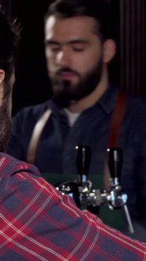 Neşeli bir erkek müşteri barda lezzetli bir zanaat birasının tadını çıkarırken kameraya ışık saçıyor. Arka planda arkadaş canlısı bir barmenin olduğu barın sıcak havası rahatlamanın ve kutlamanın davetkar atmosferini arttırıyor.