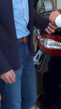 Müşteri, modern bir galeride çalışan bir satıcıdan güvenli bir şekilde araba anahtarı alır ve arka planda yeni, gösterişli araçlar bulunur. El sıkışmaları başarılı bir alışverişi sembolize ediyor, yeni bir araba almanın heyecanını vurguluyor.