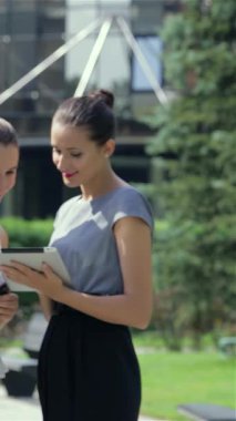 Pürüzsüz siyah takım elbiseli kendine güvenen genç bir iş kadını, açık hava ofis ortamında nişanlısı ile birlikte tabletinin üzerinde çalışıyor. Sahne profesyonelliği ve işbirliğini yayıyor, dinamik enerjiyi gösteriyor