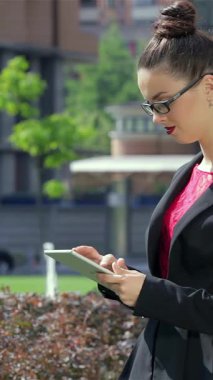 Pürüzsüz siyah takım elbiseli kendine güvenen genç bir iş kadını, açık hava ofis ortamında nişanlısı ile birlikte tabletinin üzerinde çalışıyor. Sahne profesyonelliği ve işbirliğini yayıyor, dinamik enerjiyi gösteriyor