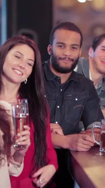 Canlı bir grup genç arkadaş, iki erkek ve iki kadın, hareketli bir barda toplanmış, neşeyle bira bardaklarını kaldırırken başparmaklarını kaldırıyorlar. Bulaşıcı gülümsemeleri ve enerjik enerjileri, neşeli bir atmosfer yaratıyor.