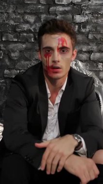 Keskin siyah bir takım elbise giymiş çarpıcı bir erkek vampir, kumlu tuğla bir duvara karşı lüks siyah bir kanepede oturuyor. Perili bakışları ve kan lekeli yüzü, ürkütücü bir Cadılar Bayramı atmosferini çağrıştırıyor. Gotik zerafetin özünü yakalamak için mükemmel.