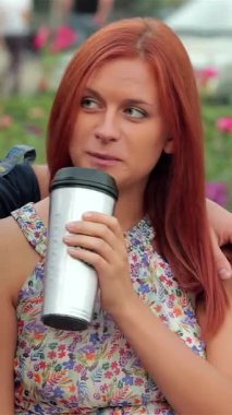 Neşeli bir çift, çiçek açan güllerle çevriliyken şık bir bardaktan kahve yudumlayarak parkta bir banka yakın otururlar. Neşeli gülümsemeleri ve samimi kucaklaşmaları canlı şehir ortamında sıcak bir sevgi ve bağlantı ortamı yaratıyor.