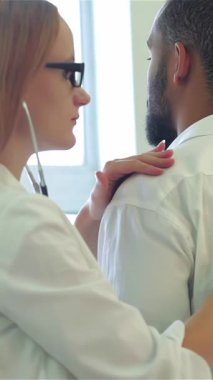 Parlak ve modern bir klinikte odaklanmış bir kadın doktor, erkek hastaları dinlemek için steteskop kullanır ve kapsamlı bir muayene sağlar. Sıcak atmosfer onların profesyonel etkileşimlerini vurguluyor. Doktor için bir reçete yazmaya hazırlanıyor.