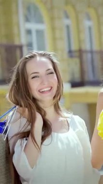 İki neşeli kız, biri parlak sarı elbiseli, diğeri beyaz bluzlu, şık alışveriş çantalarını canlı bir şehir zeminine karşı mutlu bir şekilde sergiliyorlar. Işıl ışıl gülüşleri ve kaygısız ruhları dostluğun özünü yakalar.