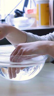 Huzurlu bir güzellik salonunda, bir kadın ellerini kristal berrak bir kase ılık suya yatırır, hoşgörülü bir maniküre hazırlanır. Renkli tırnak cilası şişeleri ve hafif ışıklandırmayla vurgulanan rahatlatıcı atmosfer mükemmel bir ışık yaratıyor.