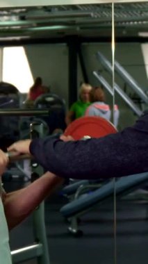 Dinamik bir spor salonu ortamında, kararlı gri saçlı yaşlı bir adam canlı kırmızı bir halteri kaldırır ve dikkatle pazularına odaklanır. Genç Kafkasyalı antrenörü yakınlarda duruyor, uzman tavsiyeleri ve teşvik sunuyor, motive edici bir atmosferi teşvik ediyor.