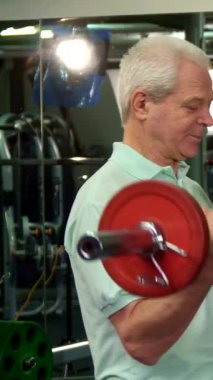 Dinamik bir spor salonu ortamında, kararlı gri saçlı son sınıf öğrencisi canlı kırmızı bir halteri kaldırır ve fitness 'e bağlılığını gösterir. Özenli genç Kafkasyalı antrenörü yakınlarda duruyor, uzman tavsiyeleri ve teşvik önerileri sunuyor, motivasyon yaratıyor.