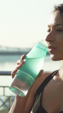 Bu yakın plan, serin sudan bir yudum alan güzel bir genç kadını yakalıyor. Işıl ışıl gülüşü, enerji veren bir egzersizin ardından yapılan susuzluğun verdiği keyfi yansıtıyor. Işıldayan şehir zeminine karşı koyduğunda, canlılığın özünü somutlaştırıyor ve