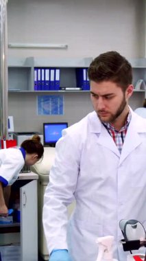 El değmemiş beyaz laboratuvar önlüğü giyen odaklanmış beyaz erkek bir bilim adamı karmaşık laboratuvar ekipmanlarını inceliyor, eldivenli eli parlak bir bilgisayar ekranının yanında geziniyor. Canlı laboratuvar çevresi enerji ile uğulduyor.