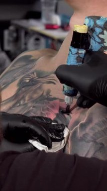 Bu çarpıcı arka planda, kendini adamış bir dövme sanatçısı bir erkek müşterinin göğsüne ustalıkla mürekkep uygular ve hikâye anlatan karmaşık vücut sanatını gözler önüne serer. Detaylara ve dinamik renklere gösterilen titiz ilgi büyüleyici bir atmosfer yaratıyor.